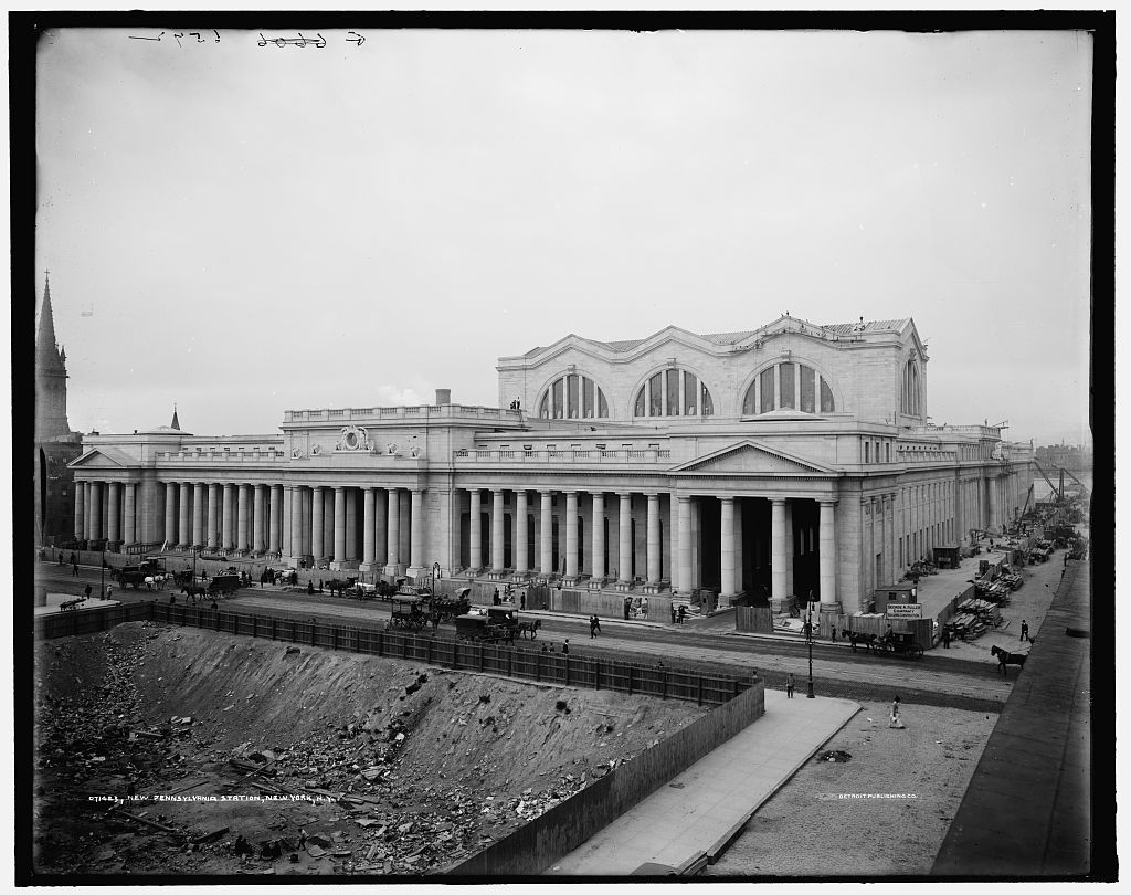 The original Pennsylvania Station around 1910: a long colonnade of granite columns and three great arched windows, horse-drawn wagons on the street, and an excavated lot in the foreground.