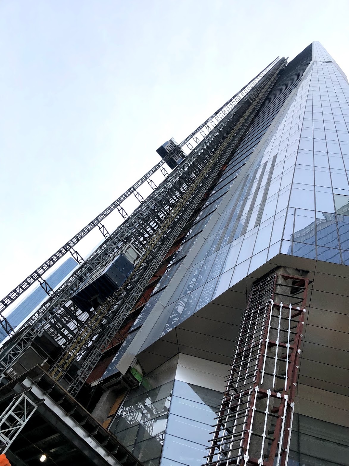 Looking straight up the face of the glass tower under construction, with an exterior hoist and steel framing climbing its side.