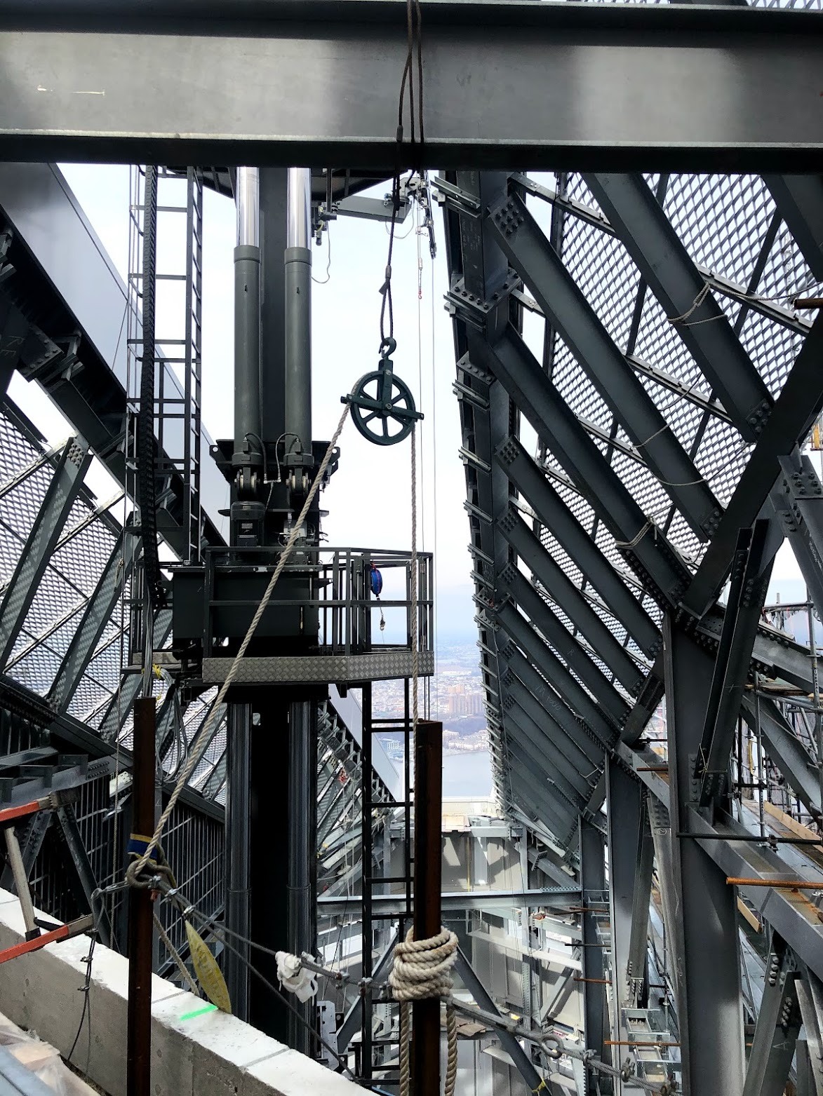 High in the tower’s crown: steel members, mesh screens, a hoist mast and a pulley, with the Hudson River visible far below.