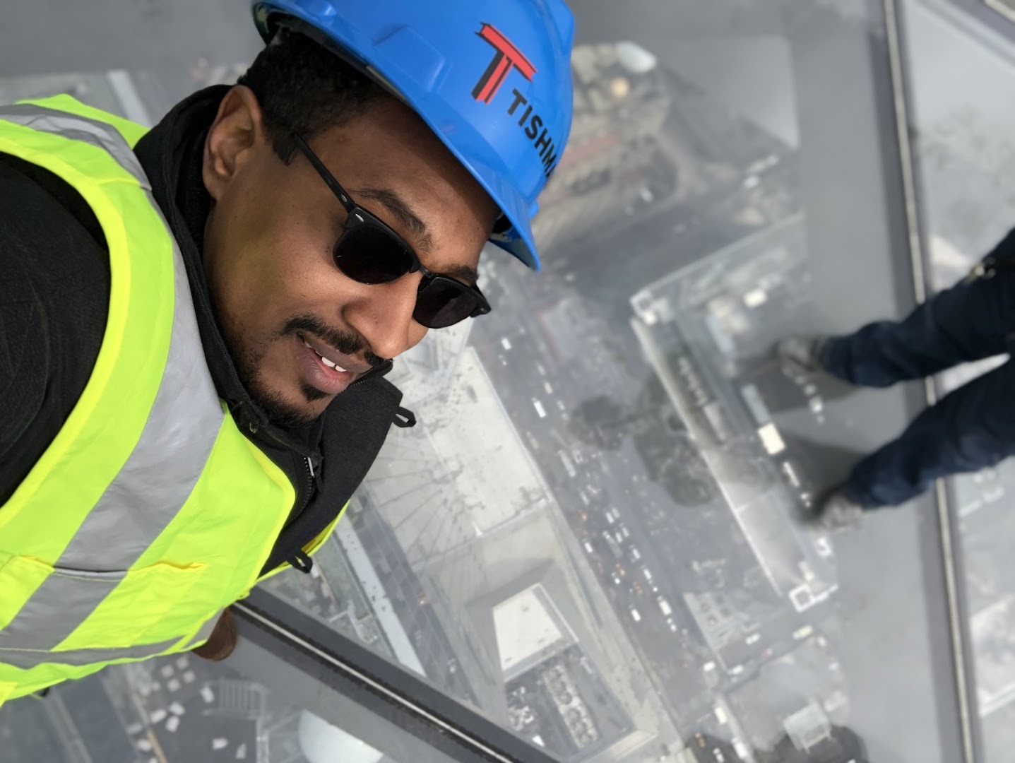 James Ward in a hard hat and safety vest taking a selfie on the glass floor of Edge, with Manhattan far below.