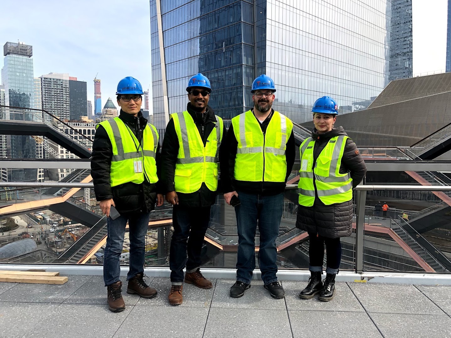 Four members of the Tishman project team, including James Ward, in hard hats and safety vests on an elevated deck at Hudson Yards, with glass towers and the Vessel’s stairways behind them.