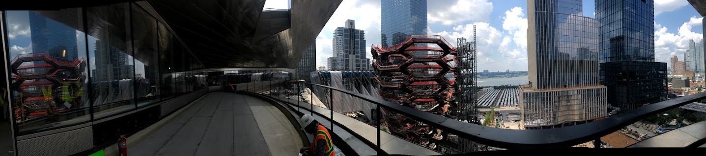 Panoramic view from an elevated walkway at Hudson Yards during construction, with the honeycomb-like Vessel, glass towers, and the Hudson River beyond.