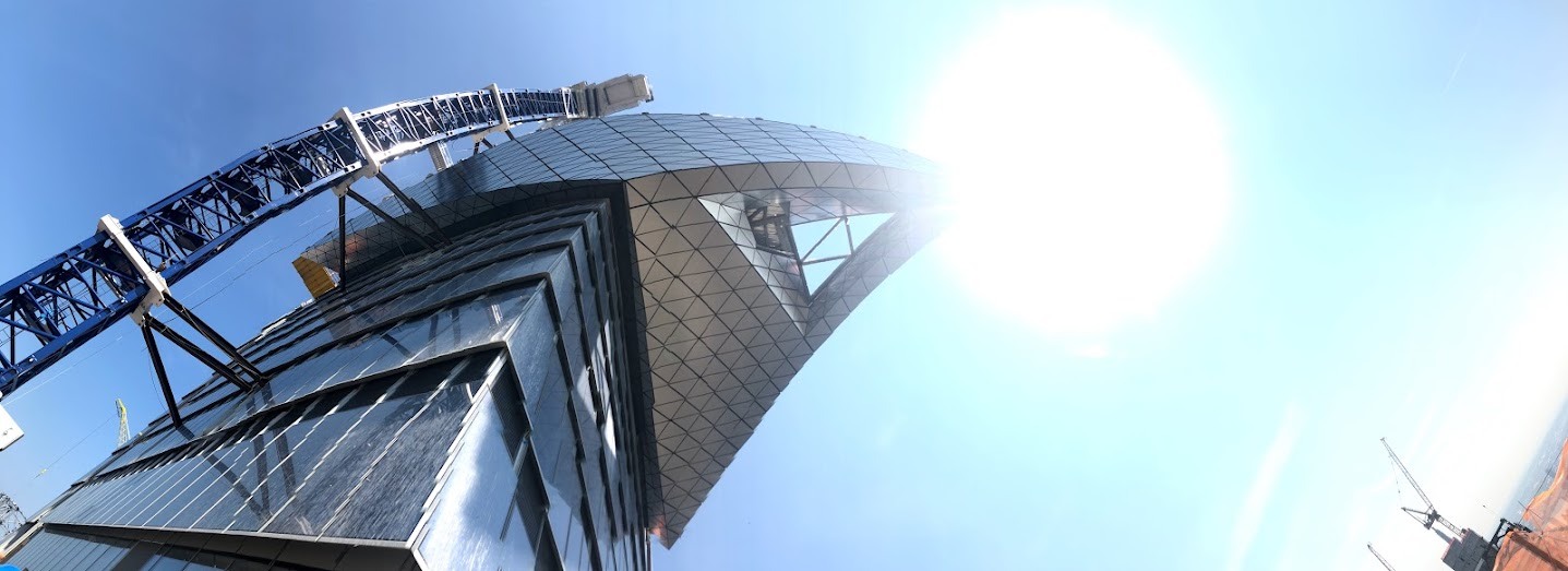 Looking up from below at Edge’s angular, triangular-paneled cantilever, with a crane boom and bright sun.