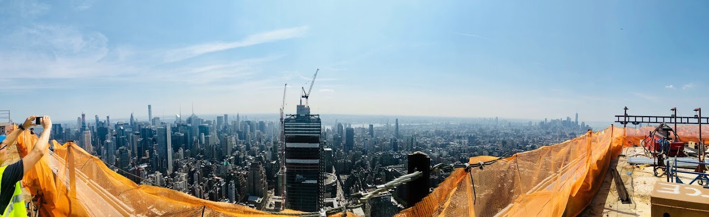 Panoramic view from the top of a tower under construction, with orange safety netting in the foreground and the Midtown Manhattan skyline stretching to the horizon.