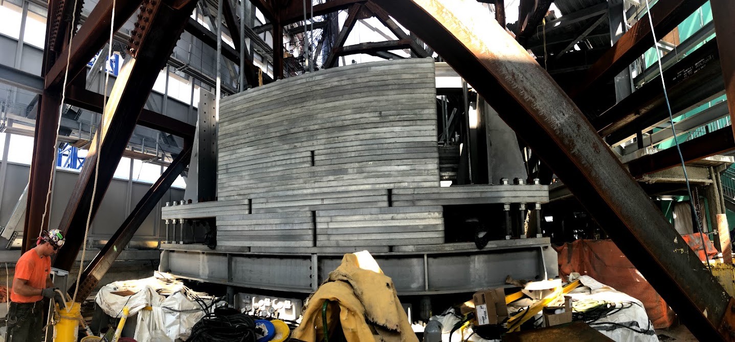 Inside a tower base: a massive layered steel assembly sits between huge diagonal steel columns while a worker in an orange shirt stands nearby.