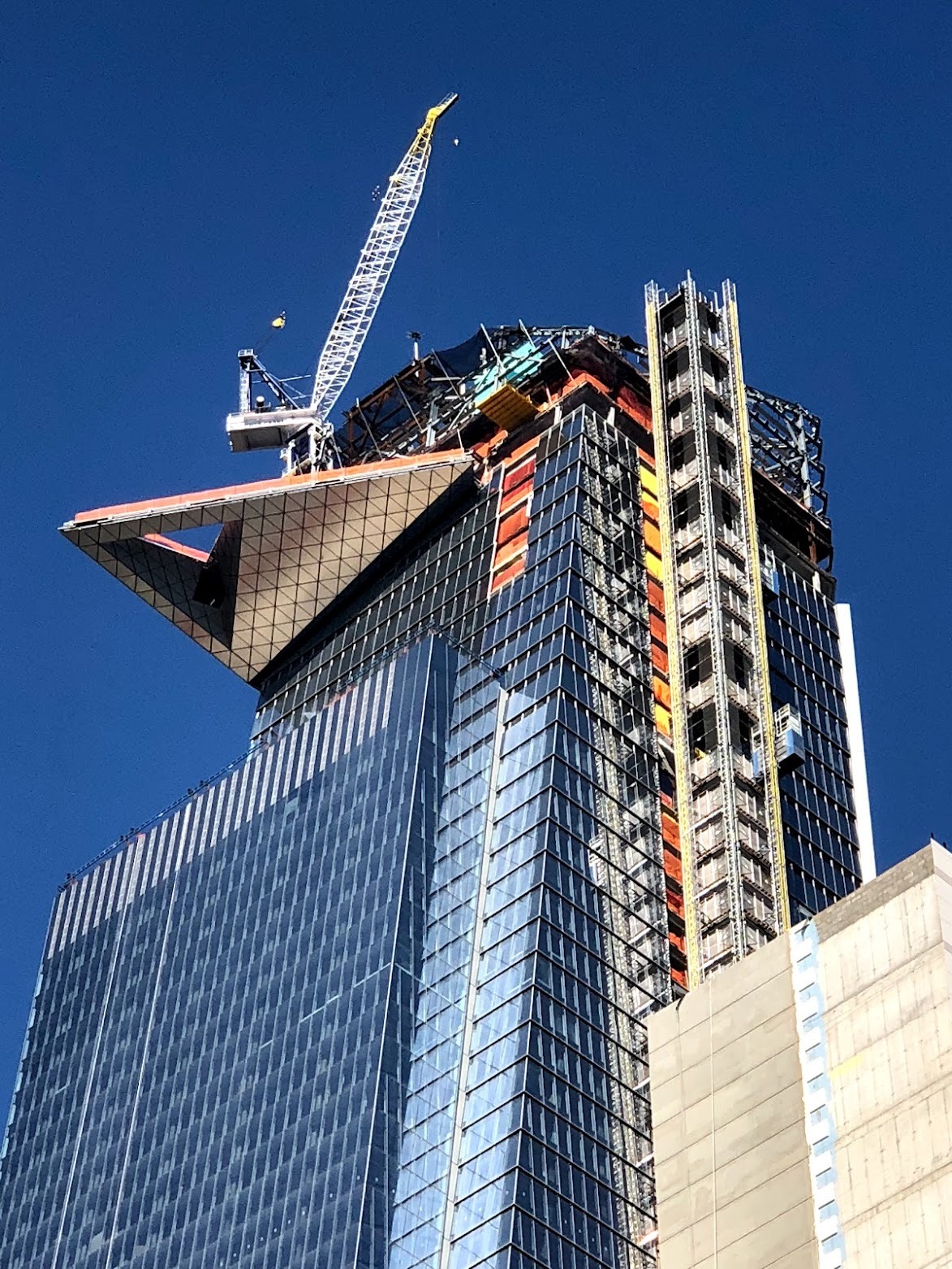 30 Hudson Yards near completion: the triangular Edge observation deck projects from the tower’s upper corner, with a crane and exterior hoist against a deep blue sky.
