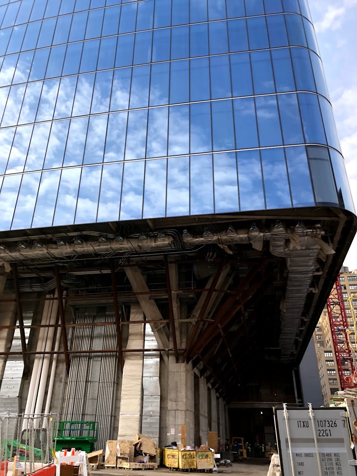 Looking up at a curved glass tower whose lower floors are still open, exposing steel framing, ductwork and concrete columns above a construction yard.