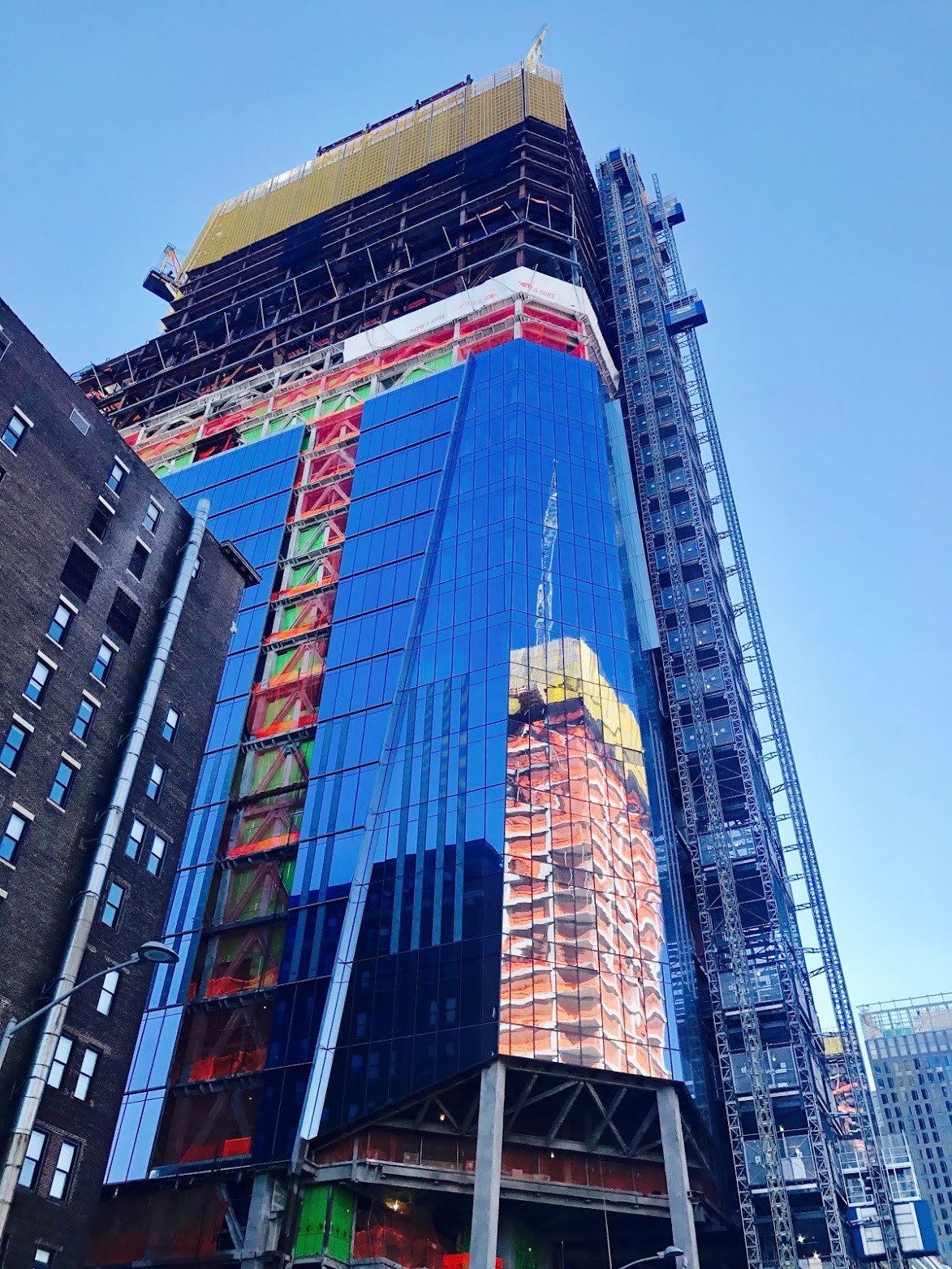A tower rising at Hudson Yards with blue glass curtain wall on its lower floors, exposed steel and coloured netting above, and an exterior hoist along one side.